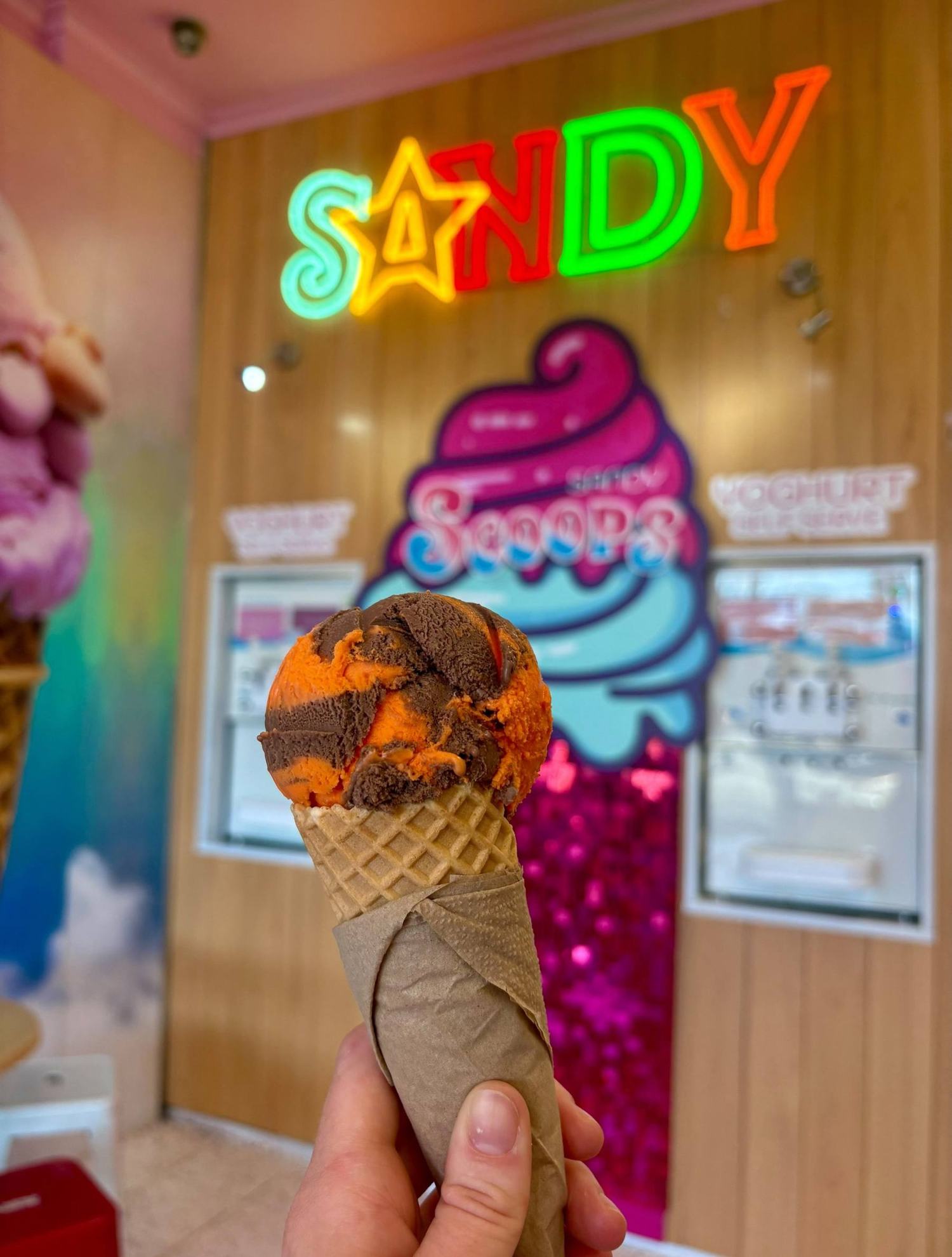 A Jaffa gelato cone held up under the neon Sandy sign and the pink swirl logo inside the shop